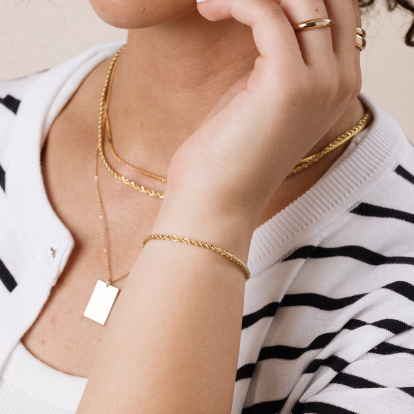 A close-up of a person wearing multiple layered gold chain necklaces, one with a rectangular pendant, with a matching gold chain bracelet and many stacked gold rings, paired with a striped white and black knit cardigan.