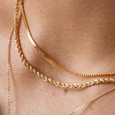 A close-up of a person wearing three gold chain necklaces, one in a rope-like texture, one with a flat braided texture, and the other a small link chain.