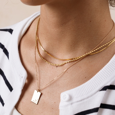 A close-up of a person wearing three layered gold necklaces, two different textured chains and one long pendant chain with a rectangular charm.