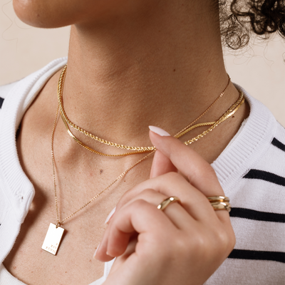 A close-up of a person wearing three layered gold necklaces, two different textured chains and one long pendant chain with a rectangular charm.
