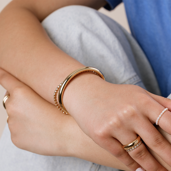 Close-up of a person's wrist wearing a two gold bracelets, one polished bangle and a tennis bracelet with circular clear gemstones, and three rings.