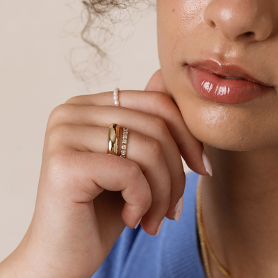 A close-up of a woman's hand resting against her cheek wearing a pearl strand ring, a wide polished gold ring and a gold ring with square bezel-set clear gemstones.