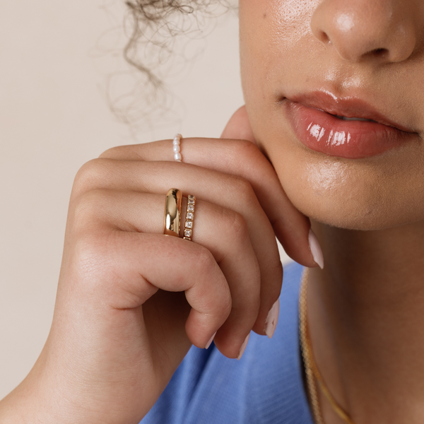 A close-up of a woman's hand resting against her cheek wearing a pearl strand ring, a wide polished gold ring and a gold ring with square bezel-set clear gemstones.