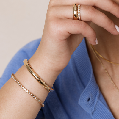 Close-up of a hand wearing gold jewelry including two bracelets, one with circular clear gemstones, two rings, and layered necklaces with a blue ribbed top.
