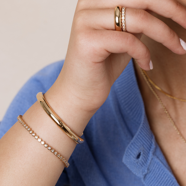 Close-up of a hand wearing gold jewelry including two bracelets, one with circular clear gemstones, two rings, and layered necklaces with a blue ribbed top.