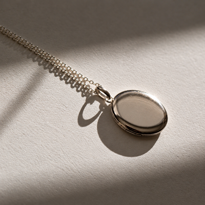 A silver oval locket necklace with a matching chain, resting on a textured beige surface under dramatic, warm sunlight with soft shadows.