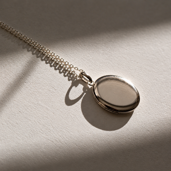 A silver oval locket necklace with a matching chain, resting on a textured beige surface under dramatic, warm sunlight with soft shadows.