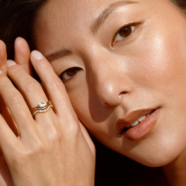 Close-up of a woman's face with one hand near her face, wearing a stack of three gold rings, one with a large tear drop shaped clear stone.