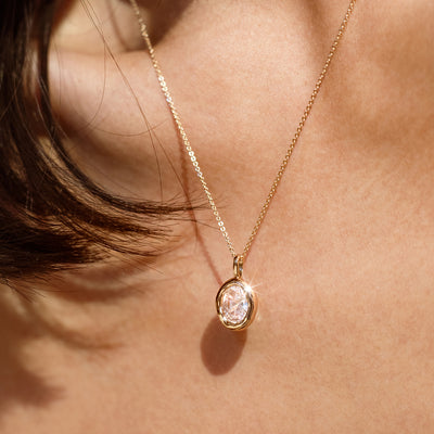 A close up lifestyle shot of a woman wearing a gold dainty chain necklace, holding a large gold bezel-set clear oval gemstone charm.