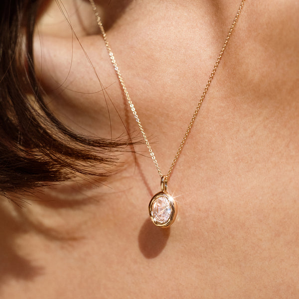 A close up lifestyle image of a person's neck wearing a gold dainty chain necklace with a large gold bezel-set clear oval gemstone charm.