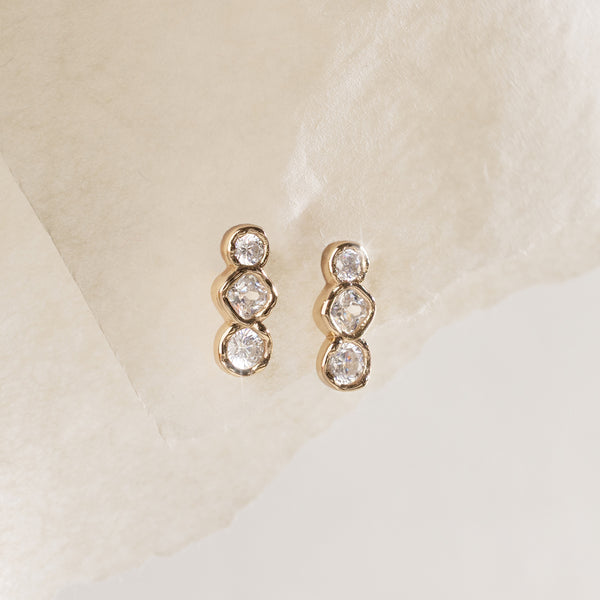 A pair of gold studs with a trio of rounded clear bezel-set gemstones, set against a white textured background.