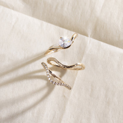 A yellow gold wavy ring trio set with a pear-cut clear center stone one, a textured, polished band second, and a row of small clear stones along the third band, shown on a light beige background.