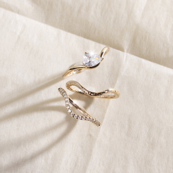A yellow gold wavy ring trio set with a pear-cut clear center stone one, a textured, polished band second, and a row of small clear stones along the third band, shown on a light beige background.