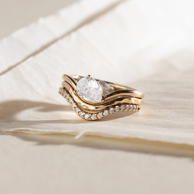 A yellow gold wavy ring trio set with a pear-cut clear center stone one, a textured, polished band second, and a row of small clear stones along the third band, shown on a light beige background.