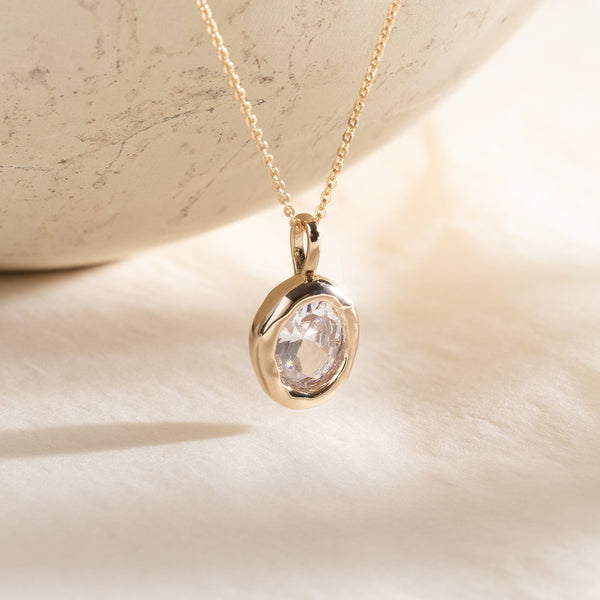 A dainty gold chain necklace with a large gold bezel-set oval clear gemstone, dangling over a textured bowl on a beige background.