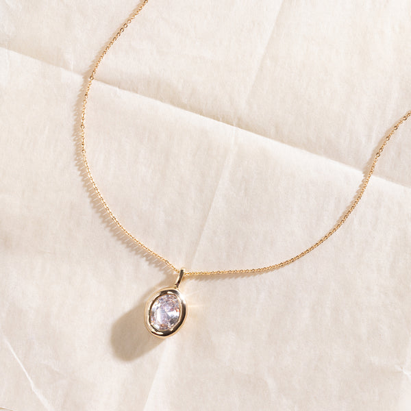A dainty gold chain pendant necklace with a large oval bezel-set clear gemstone charm, set against a textured white background.