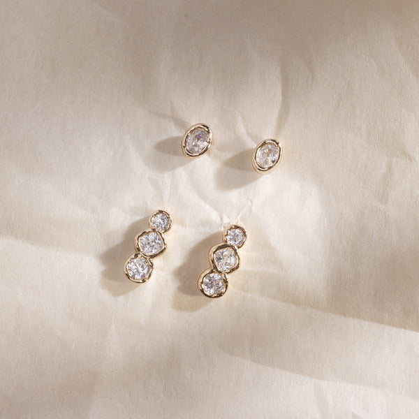 Two pairs of gold stud earrings, one with one set of clear oval bezel-set gemstones, and the other with a trio of rounded clear bezel-set gemstones, set against a textured white background with shadows.