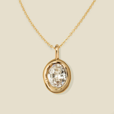 A gold necklace with a large bezel-set oval clear gemstone pendant, displayed on a light beige background.