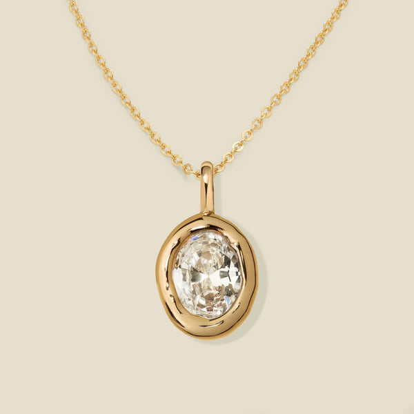 A gold necklace with a large bezel-set oval clear gemstone pendant, displayed on a light beige background.