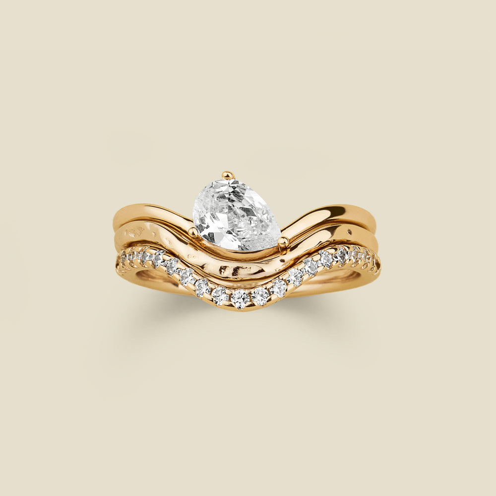 A yellow gold wavy ring trio set with a pear-cut clear center stone one, a textured, polished band second, and a row of small clear stones along the third band, shown on a light beige background.