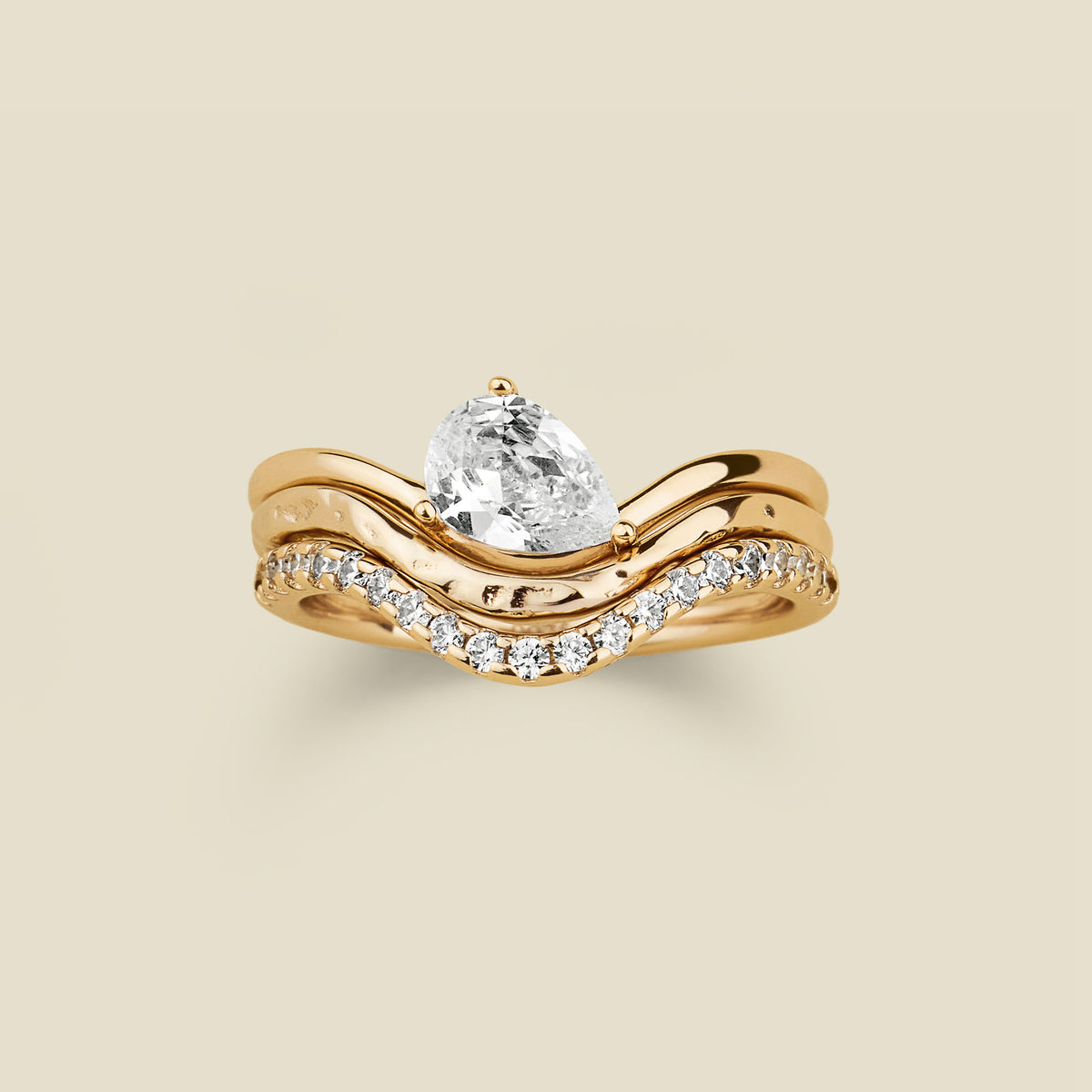 A yellow gold wavy ring trio set with a pear-cut clear center stone one, a textured, polished band second, and a row of small clear stones along the third band, shown on a light beige background.