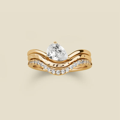 A yellow gold wavy ring trio set with a pear-cut clear center stone one, a textured, polished band second, and a row of small clear stones along the third band, shown on a light beige background.