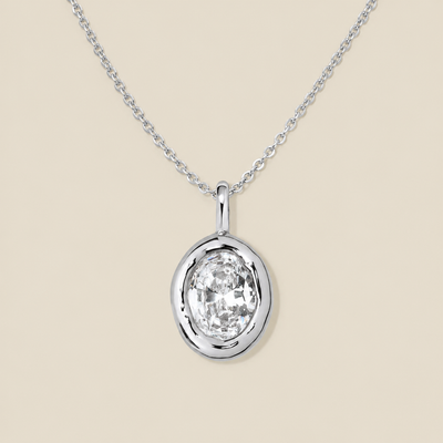 A large oval bezel-set clear gemstone charm on a dainty silver chain, set against a solid beige background.