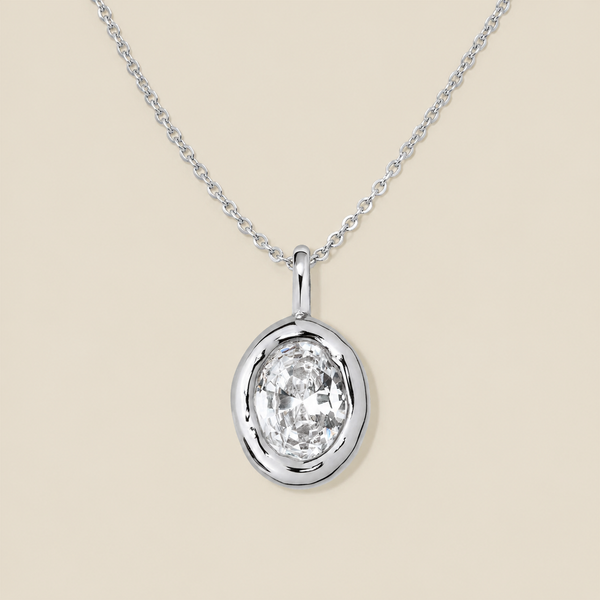 A large oval bezel-set clear gemstone charm on a dainty silver chain, set against a solid beige background.