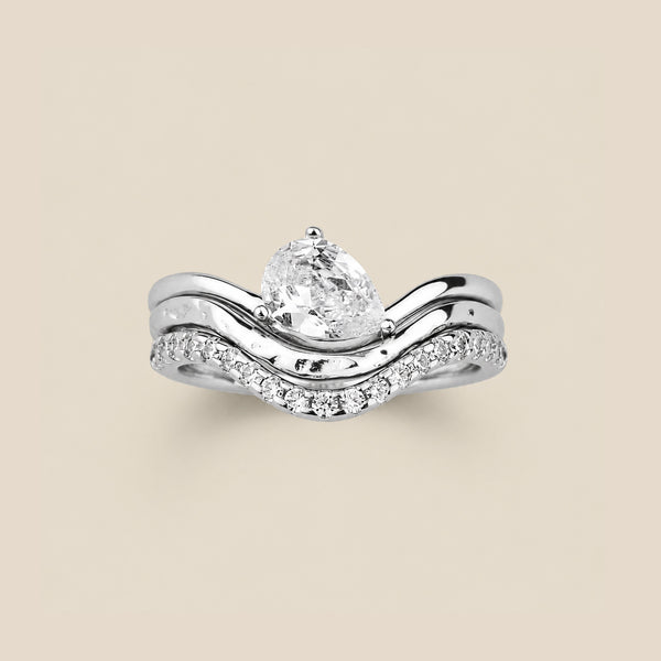 A silver ring trio set stacked together, with all the rings dipping down in the front. The bottom ring has small clear stones set along the front, the middle ring has a textured pattern, and the top ring features a large tear-drop prong set clear gemstone turned to one side. The ring stack is set on a solid beige background.