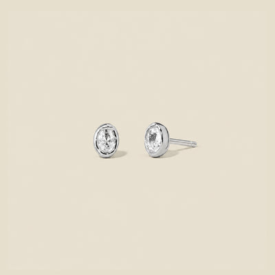 Pair of silver stud earrings with clear oval bezel-set gemstones on a beige background.