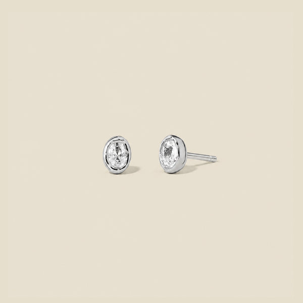 Pair of silver stud earrings with clear oval bezel-set gemstones on a beige background.