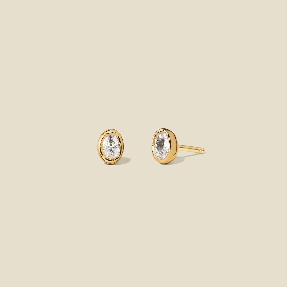 Pair of gold stud earrings with clear bezel-set oval gemstones on a beige background