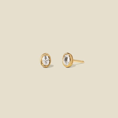 Pair of gold stud earrings with clear bezel-set oval gemstones on a beige background