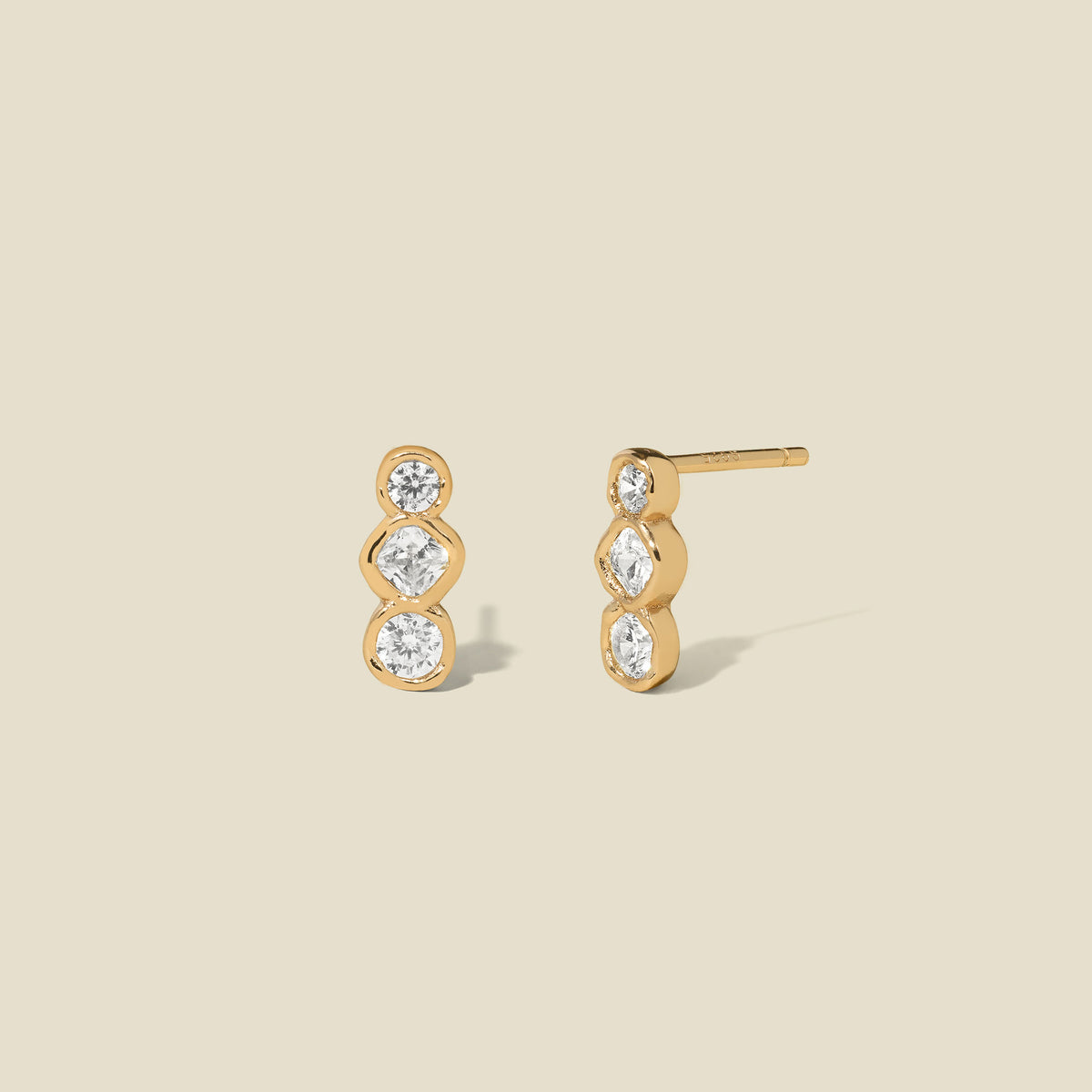 Gold stud earrings with three clear bezel-set vertical gemstones on a beige background