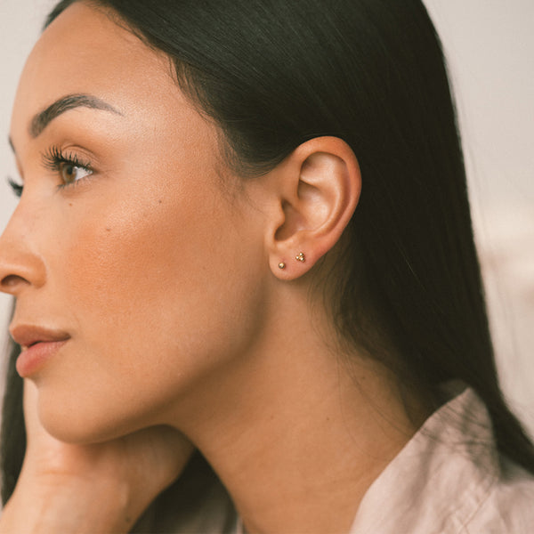 Close-up of a woman's ear wearing two small gold stud earrings, with her face in profile against a soft light background.