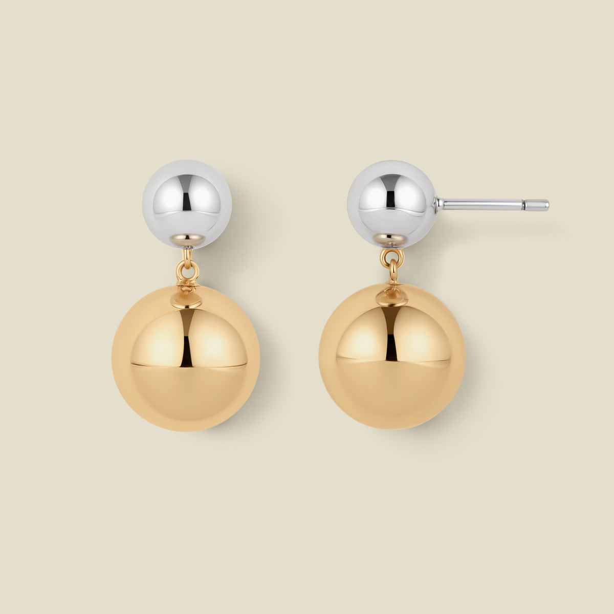 A pair of minimalist two-tone ball drop earrings on a solid light beige background. Each earring features a polished silver-tone ball stud connected by a small gold chain to a larger, polished gold-tone ball dangle.