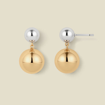 A pair of minimalist two-tone ball drop earrings on a solid light beige background. Each earring features a polished silver-tone ball stud connected by a small gold chain to a larger, polished gold-tone ball dangle.