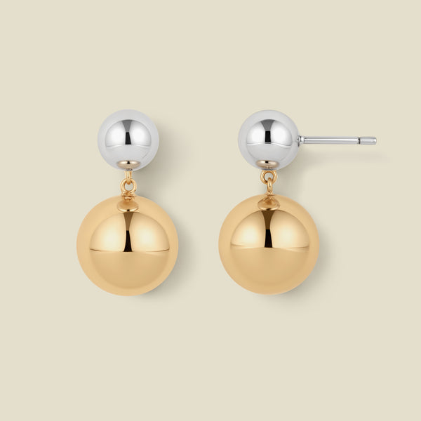 A pair of minimalist two-tone ball drop earrings on a solid light beige background. Each earring features a polished silver-tone ball stud connected by a small gold chain to a larger, polished gold-tone ball dangle.