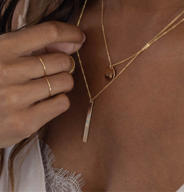 A close-up lifestyle shot of a model wearing a layered gold necklace set, featuring a delicate chain with a flat rectangular bar pendant and a second chain with a small circular disc pendant with rainbow design engraved.