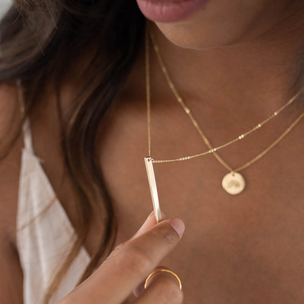 A close-up lifestyle shot of a model wearing layered gold necklaces, including a delicate bar necklace being held by a hand and a gold disc pendant necklace.