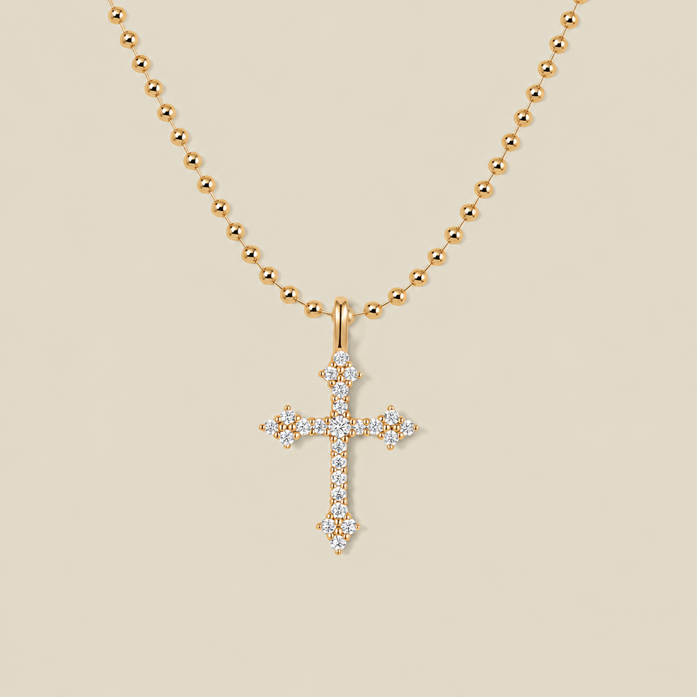 A delicate gold bead chain necklace with a cross charm covered in clear stones against a solid beige background.