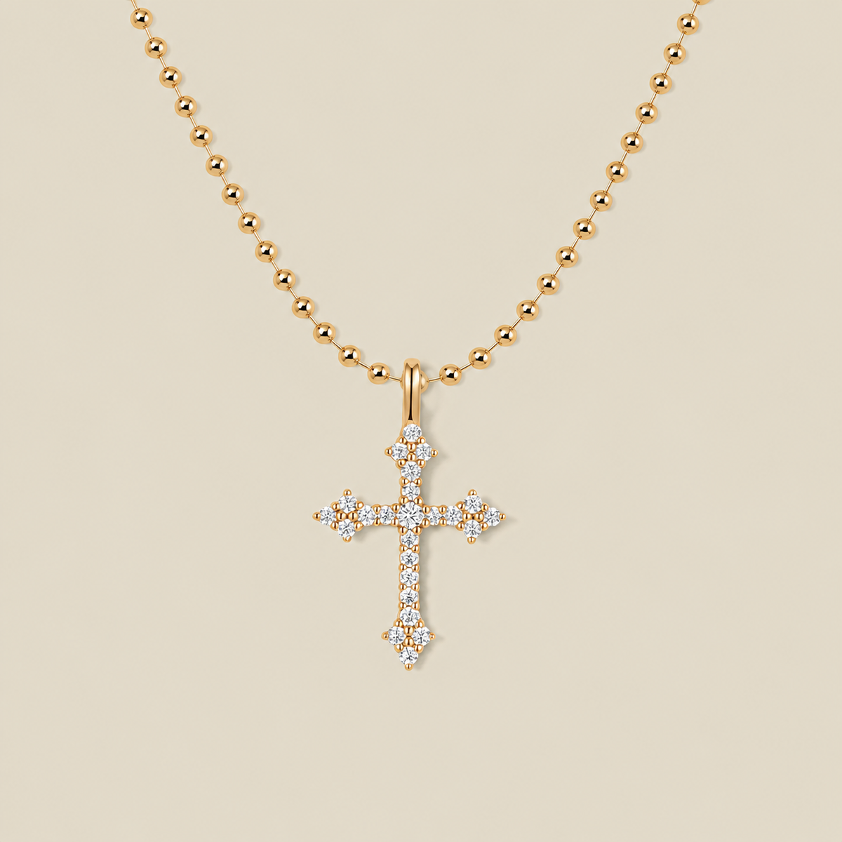 A delicate gold bead chain necklace with a cross charm covered in clear stones against a solid beige background.