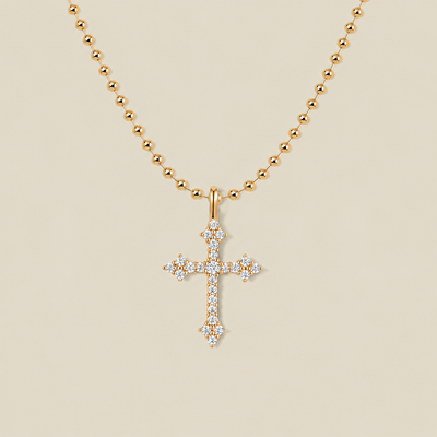 A delicate gold bead chain necklace with a cross charm covered in clear stones against a solid beige background.