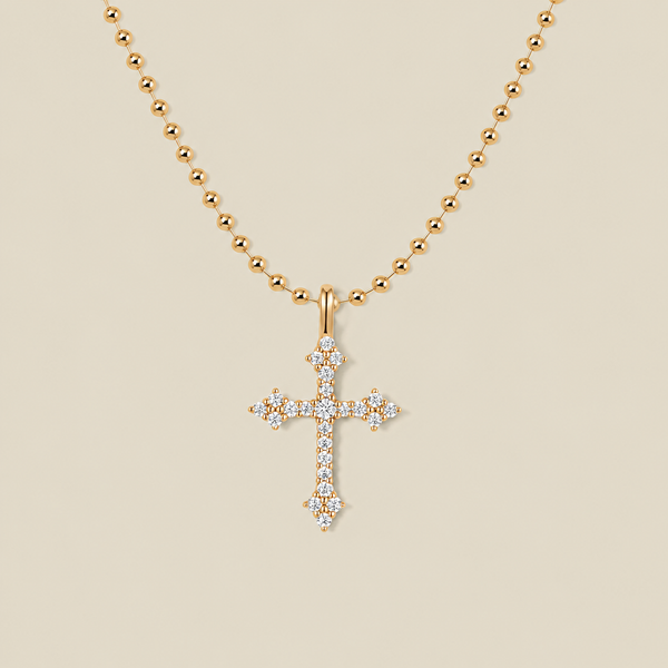 A delicate gold bead chain necklace with a cross charm covered in clear stones against a solid beige background.