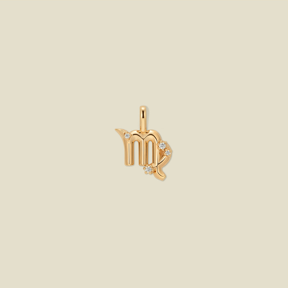 A gold pendant shaped like the Virgo zodiac symbol, featuring three small clear gemstones, centered on a solid light beige background.
