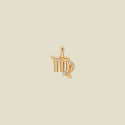 A gold pendant shaped like the Virgo zodiac symbol, featuring three small clear gemstones, centered on a solid light beige background.