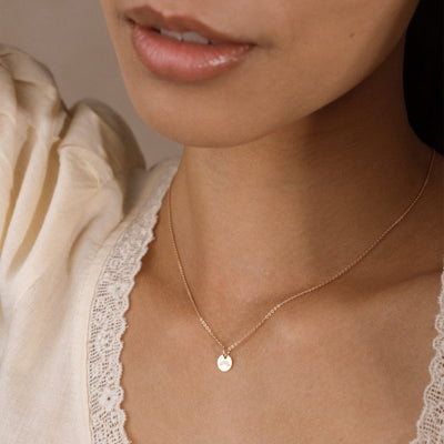 A close-up of a model wearing a delicate gold chain necklace with a small, flat circular disc pendant with an engraved rainbow. The model is wearing a cream-colored lace-trimmed top, and the background is a soft, neutral beige.