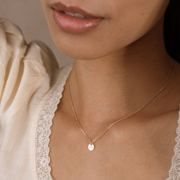 A close-up of a model wearing a delicate gold chain necklace with a small, flat circular disc pendant with an engraved rainbow. The model is wearing a cream-colored lace-trimmed top, and the background is a soft, neutral beige.