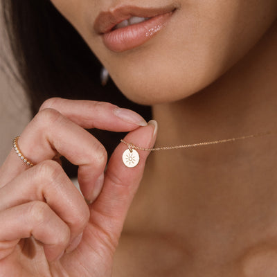 A close-up lifestyle shot of a model holding a delicate gold chain necklace with a small round disc charm engraved with a sun pattern. The model's hand, lips, and neck are visible against a warm, softly lit background.