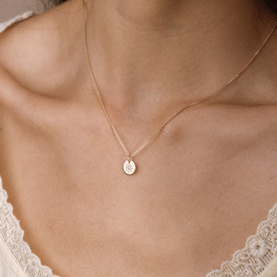 A delicate gold chain necklace with a small circular disc pendant featuring an engraved sun design, shown worn on a model's neck.